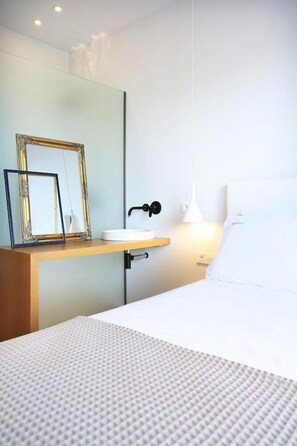 Hotelkamer met een bed, nachtkastje met spiegel en fotolijst, bedlamp en een wit kussen.