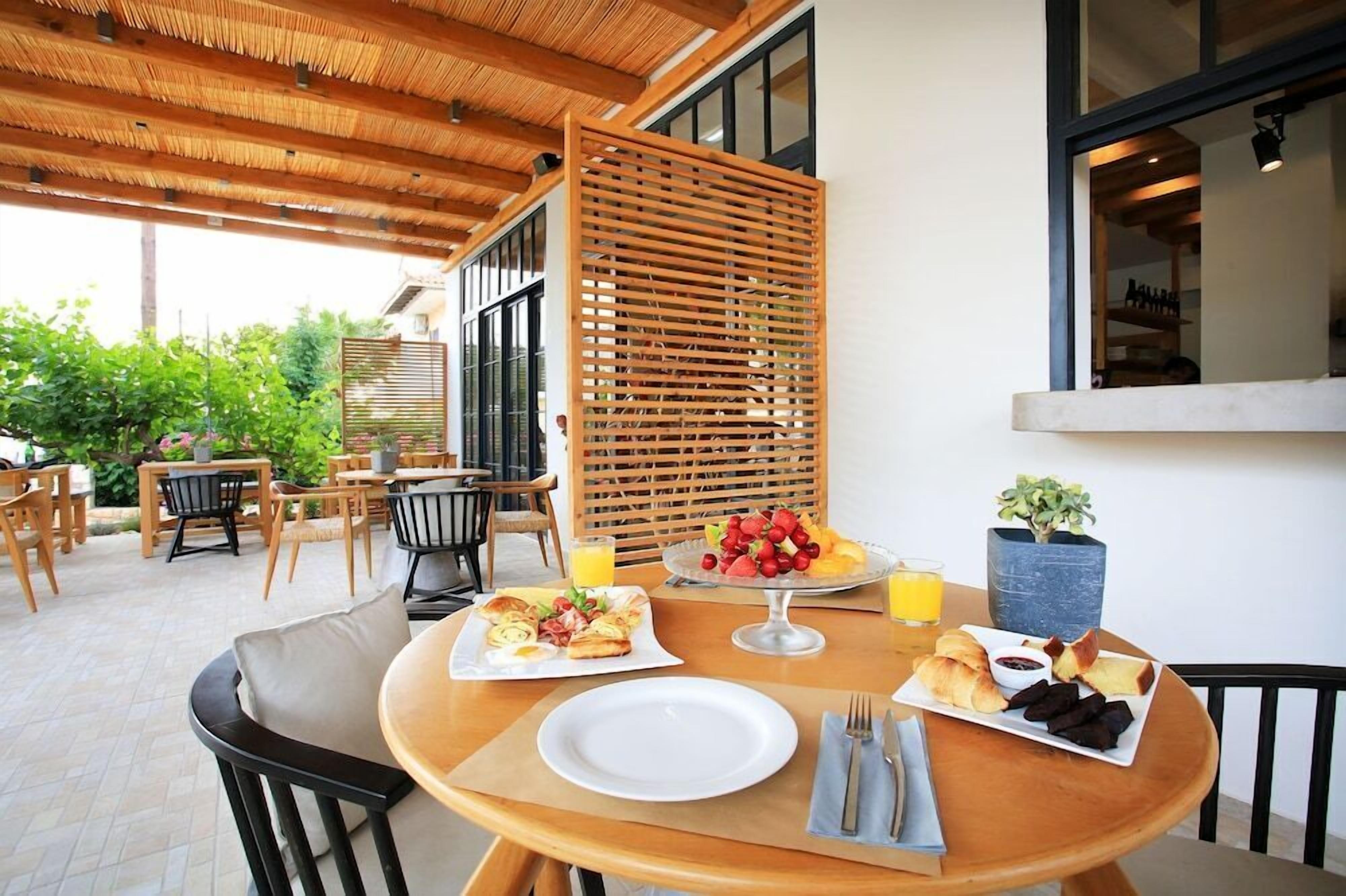 An outdoor dining area with wooden tables and chairs, a thatched roof, and a view of greenery.