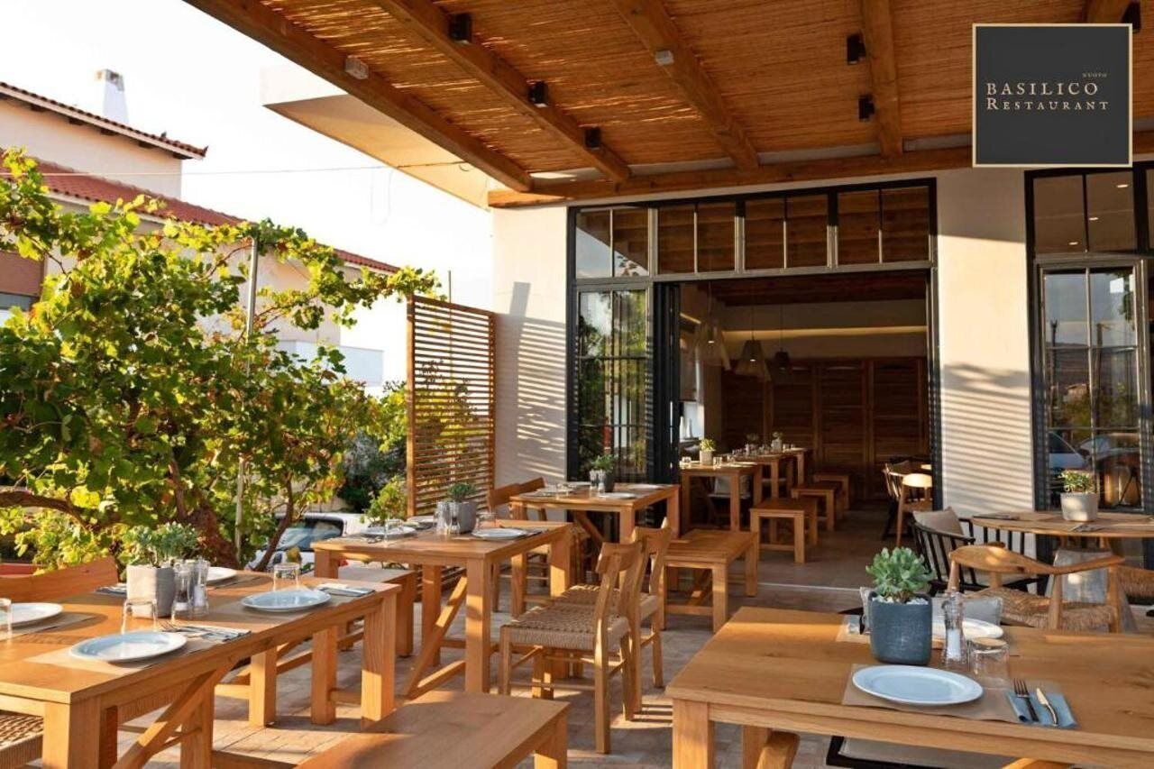Un área de comedor al aire libre con mesas y sillas de madera, un letrero para el Restaurante Basilico y vistas del interior a través de una puerta de cristal.