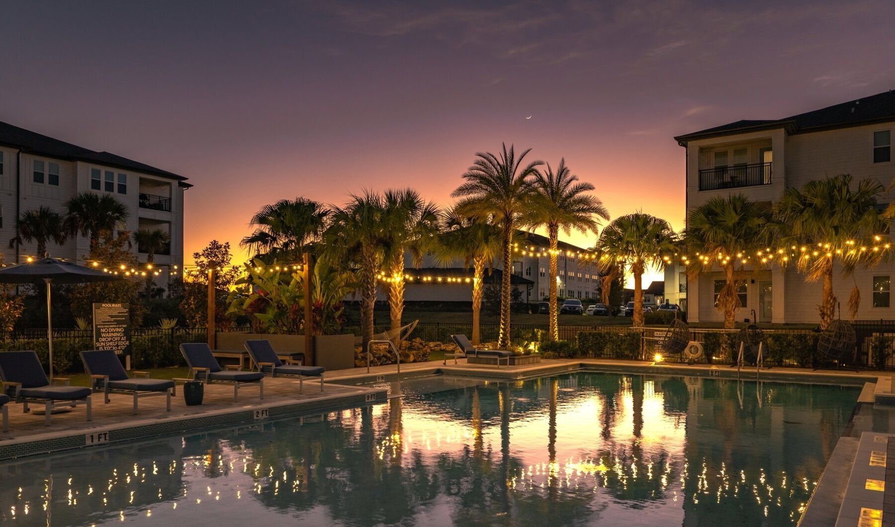 A swimming pool with lounge chairs and palm trees, surrounded by buildings during sunset.