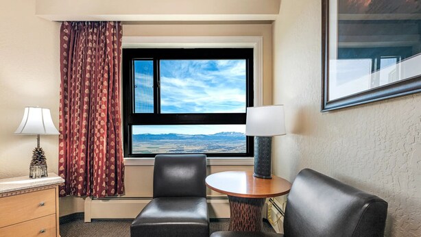 A hotel room with a bed, a desk, a chair, a lamp, a window with a mountain view, and a framed picture on the wall.