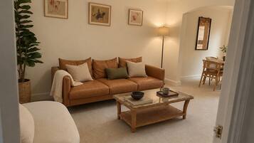 A living room with a brown leather sofa, a glass coffee table, and a large floor lamp. There are framed artworks on the wall and a potted plant.