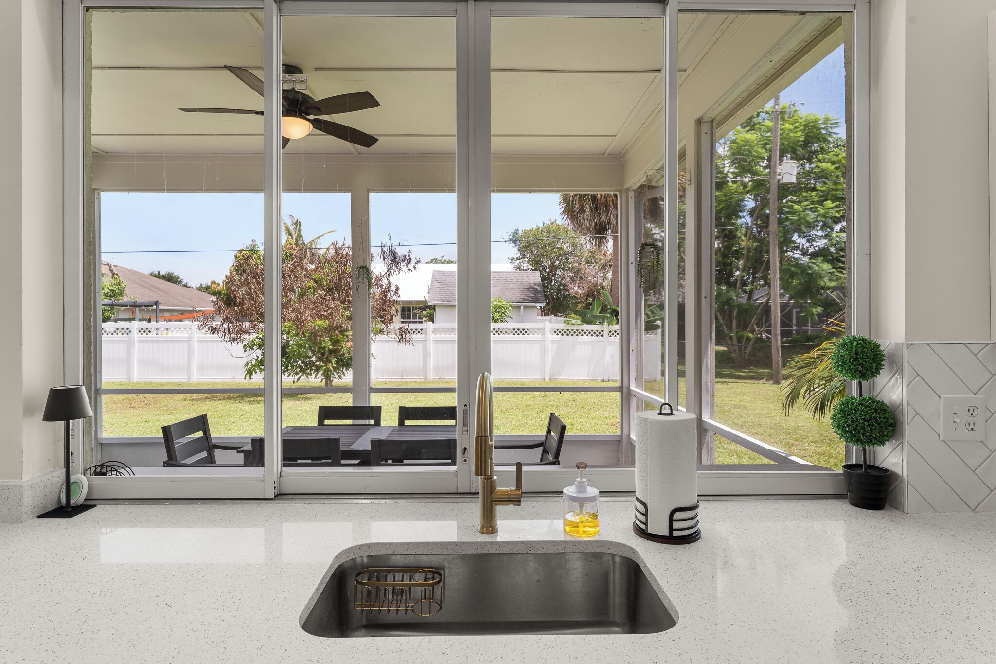 A kitchen with a stainless steel sink, a ceiling fan, and a view of a backyard with a patio and a white fence.