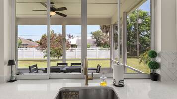 A kitchen with a stainless steel sink, a ceiling fan, and a view of a backyard with a patio and a white fence.