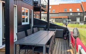 Une terrasse extérieure avec une table et des chaises, un véhicule rouge et, au fond, un bâtiment dont le toit est recouvert de tuiles orange.