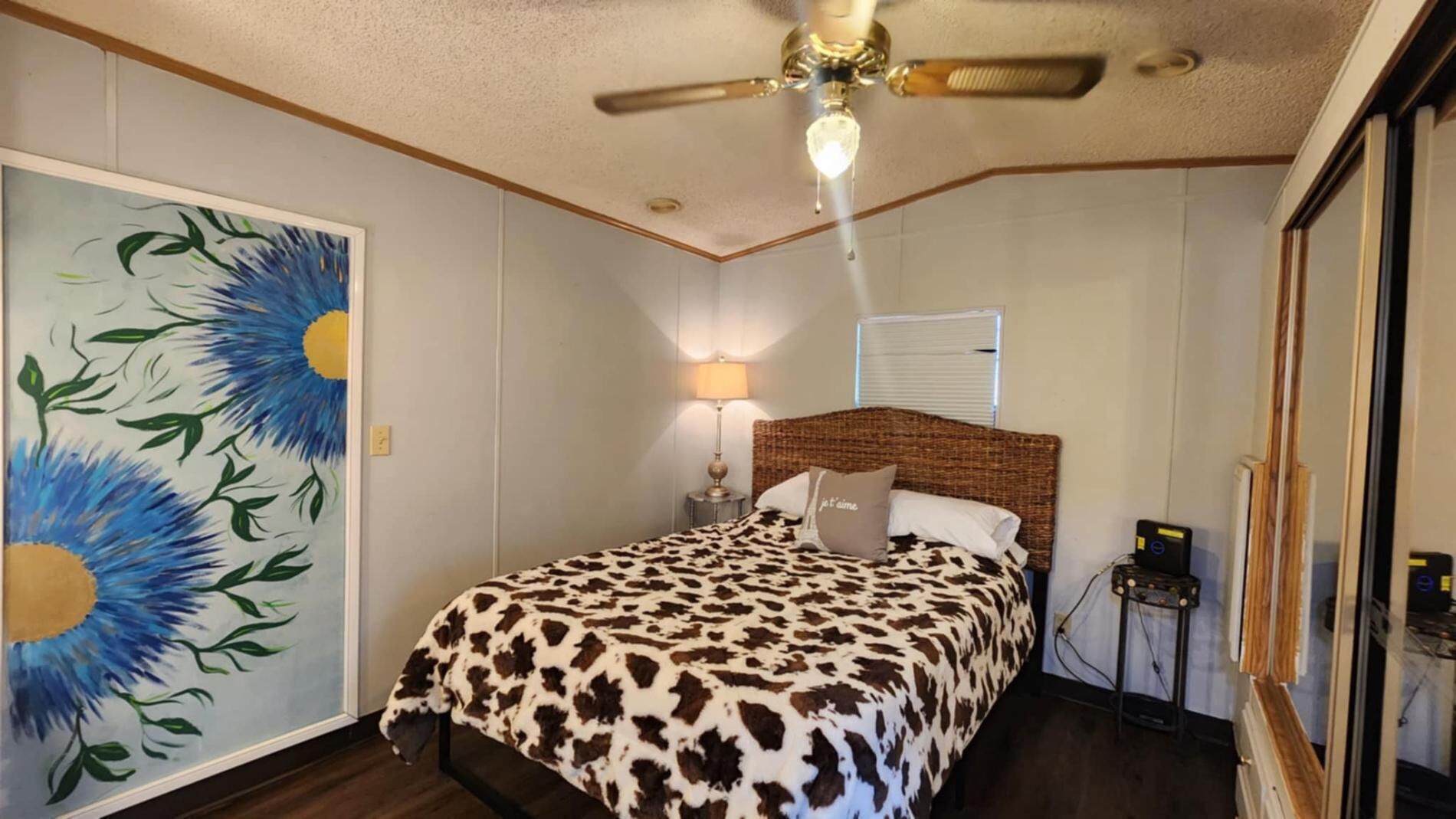 A bedroom with a bed, a ceiling fan, a nightstand, a lamp, and a painting on the wall.