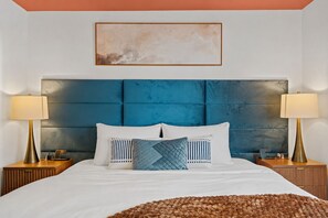 Ein Bett mit dunkelblau gepolstertem Kopfteil, zwei Nachttische mit Lampen und ein gerahmtes abstraktes Gemälde darüber.