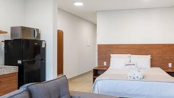 Quarto de hotel com uma cama, cabeceira de madeira, pequena escrivaninha com um refrigerador e um sofá cinzento.