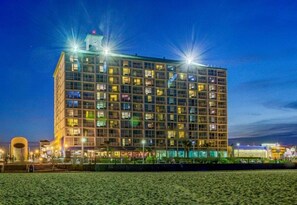 Um hotel de vários andares com design moderno, destacando uma torre do relógio imponente e diversas janelas iluminadas.