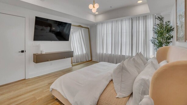 A modern bedroom with a bed, a TV mounted on the wall, a bedside table, and a large window with sheer curtains. - Modern Loft/Walk To Stadiums/Free Parking! (Atlanta)