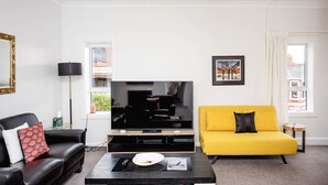 Een woonkamer met een flatscreen televisie, een gele bank, een zwarte leren bank en een salontafel.