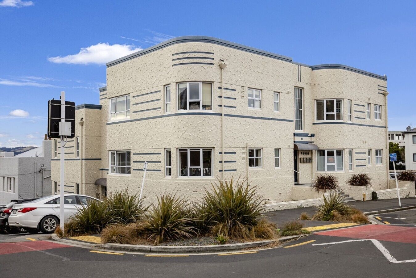 A multi-story building with a textured facade, a visible ground floor entrance, and a parking area with a white car.