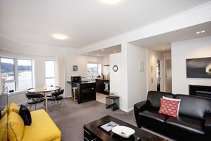 A modern living room with a sofa, yellow ottoman, and a coffee table. There is a dining area with a table and chairs, and a kitchen area with a microwave and sink. - The Deco View, Warm Art Deco Apartment with Garage (Dunedin)