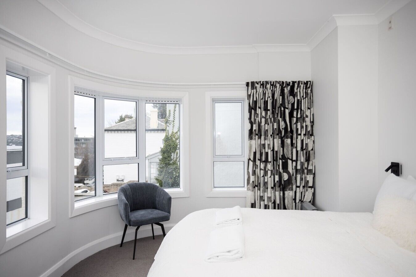 A hotel room with a large bed, a chair, a window with a view, and a curtain with a geometric pattern.