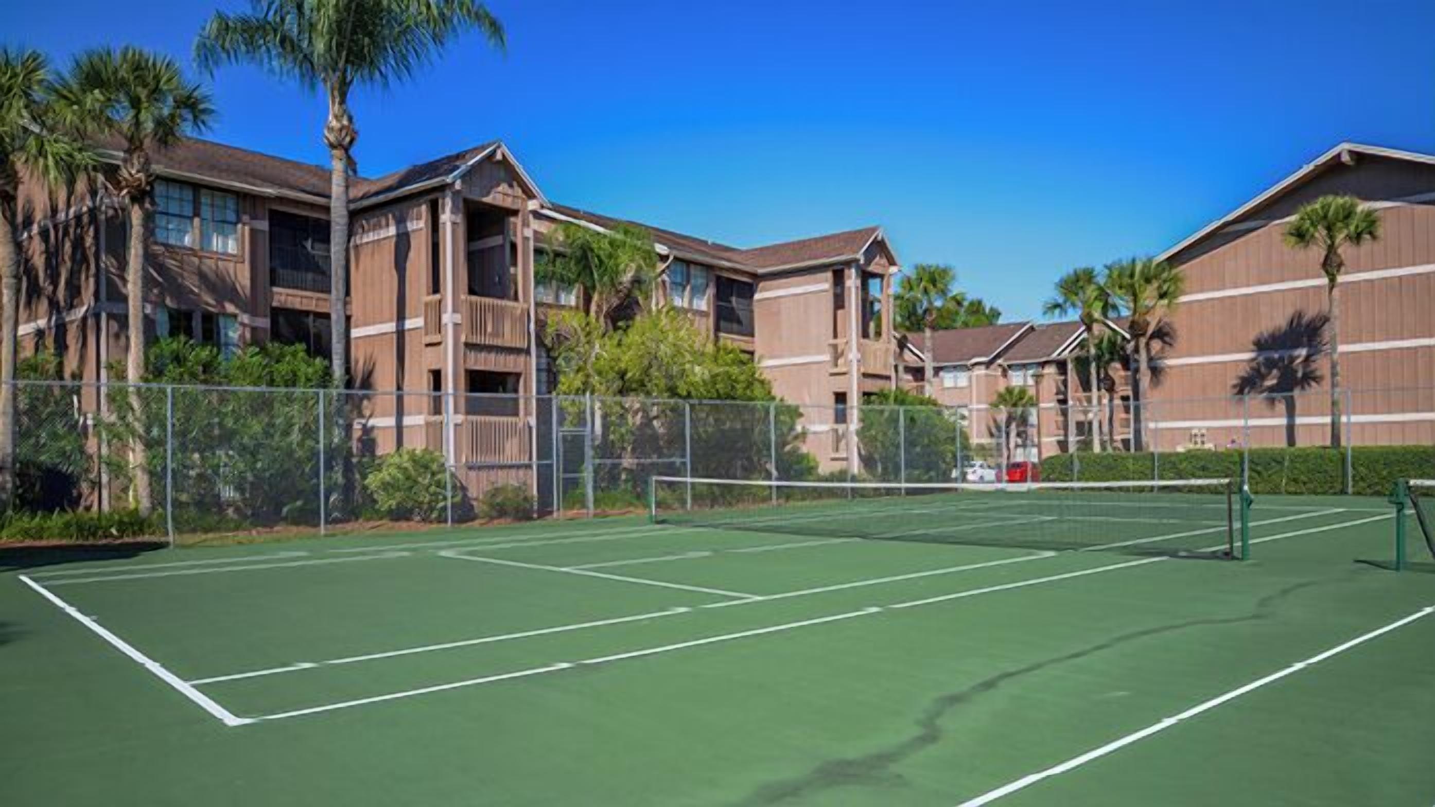 Una cancha de tenis con red, rodeada por una cerca y contigua a un edificio con varios balcones y palmeras.