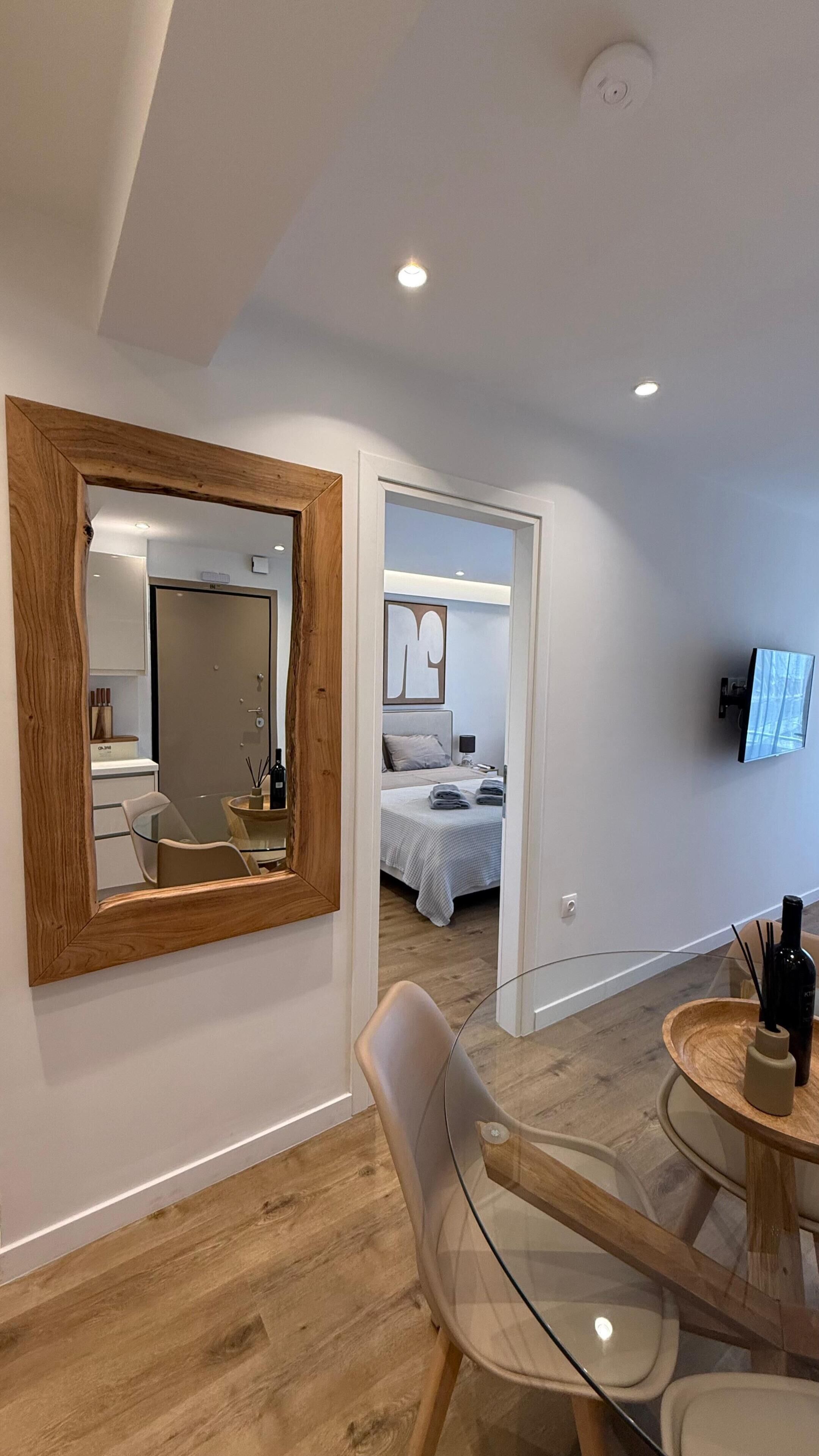 A modern hotel room with a large mirror, a glass table, a bed with a grey bedspread, and a television mounted on the wall.