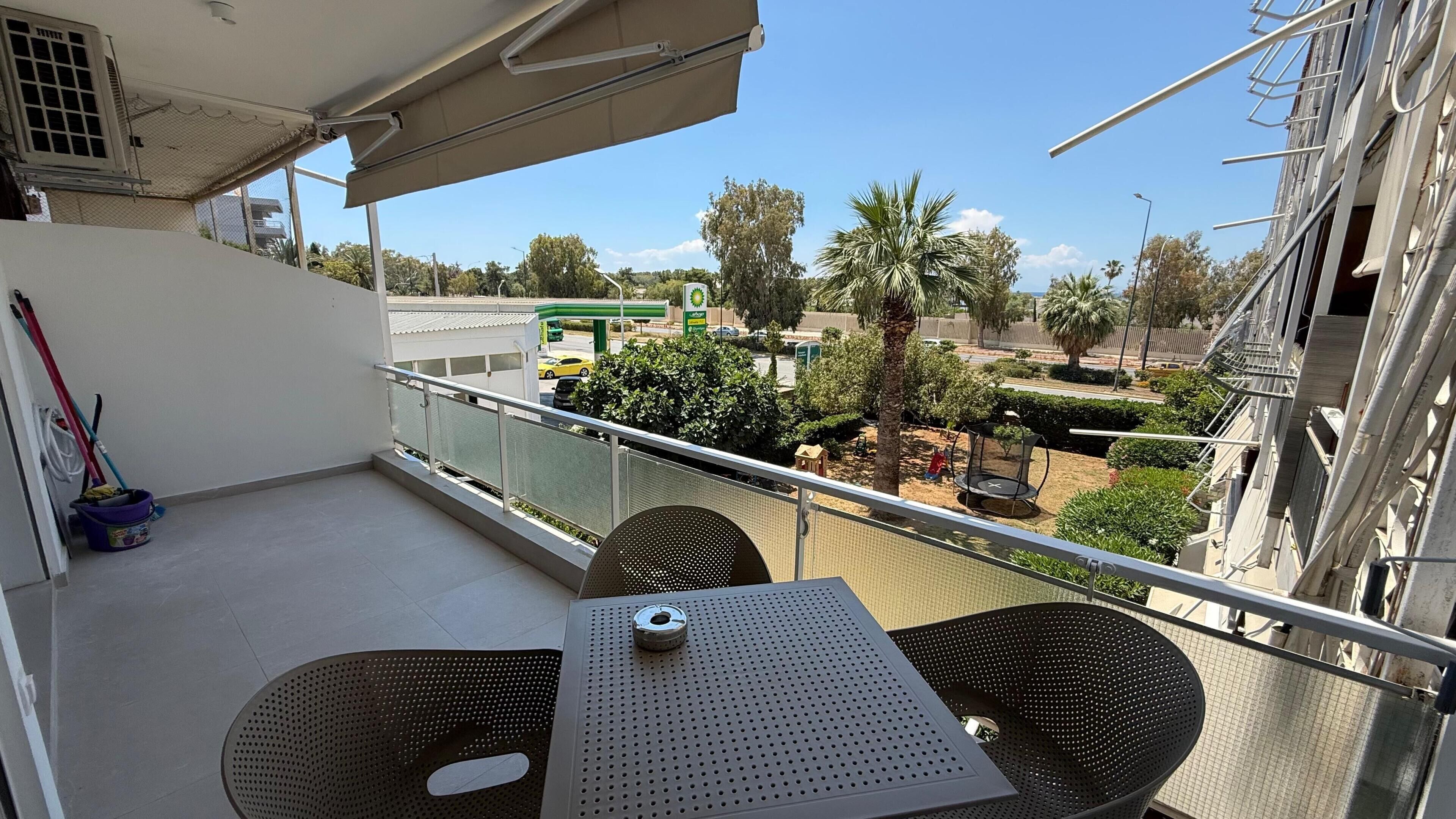 A balcony with a table and chairs, a view of a parking lot, and a green gas station in the background.
