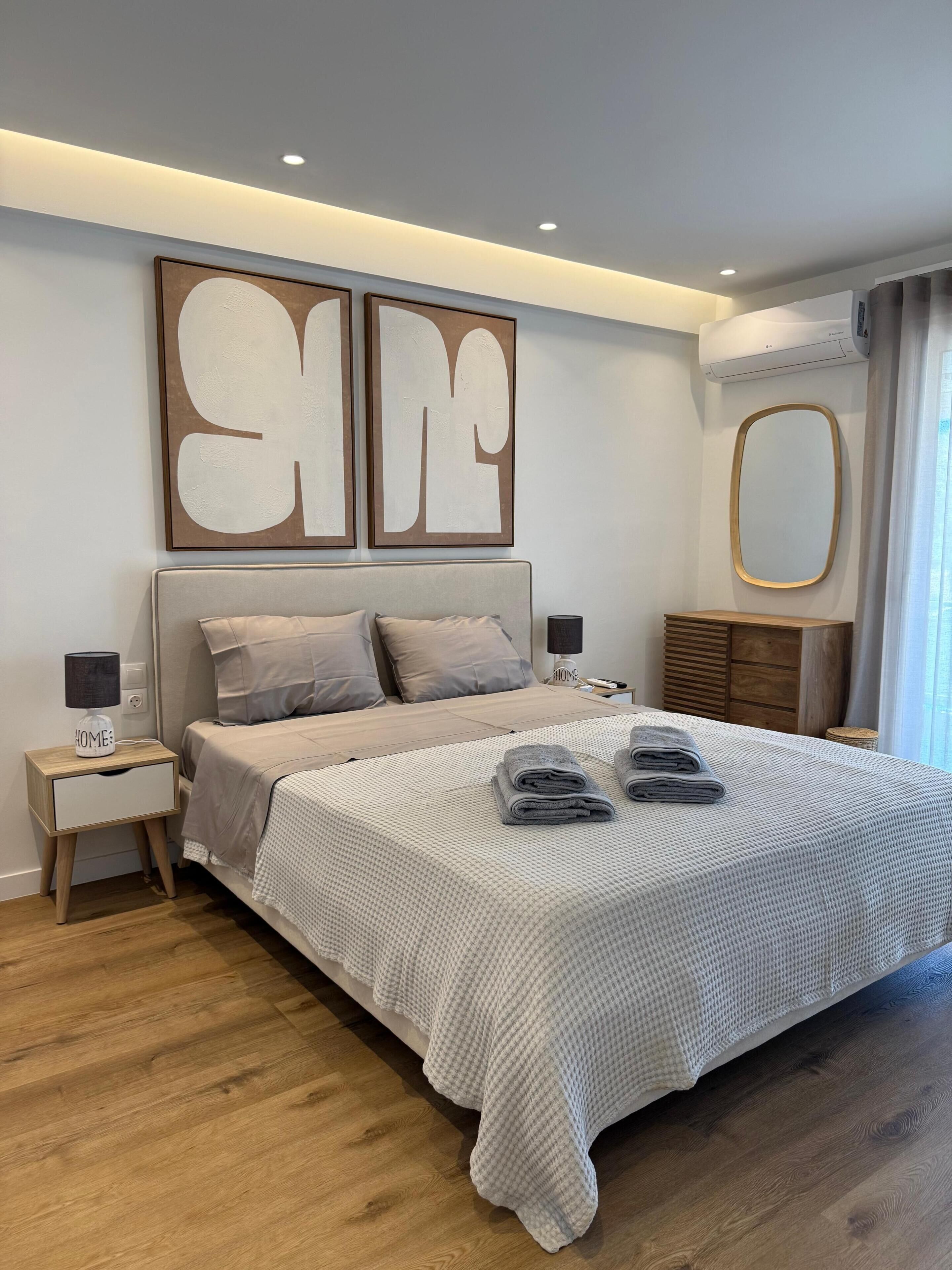 A modern bedroom with a large bed, bedside tables, a mirror, and a wall-mounted air conditioner.