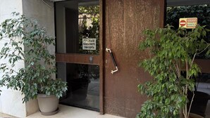 Entrada de um edifício com porta marrom, janela de vidro e uma placa em grego. Há dois vasos de plantas de cada lado da porta.