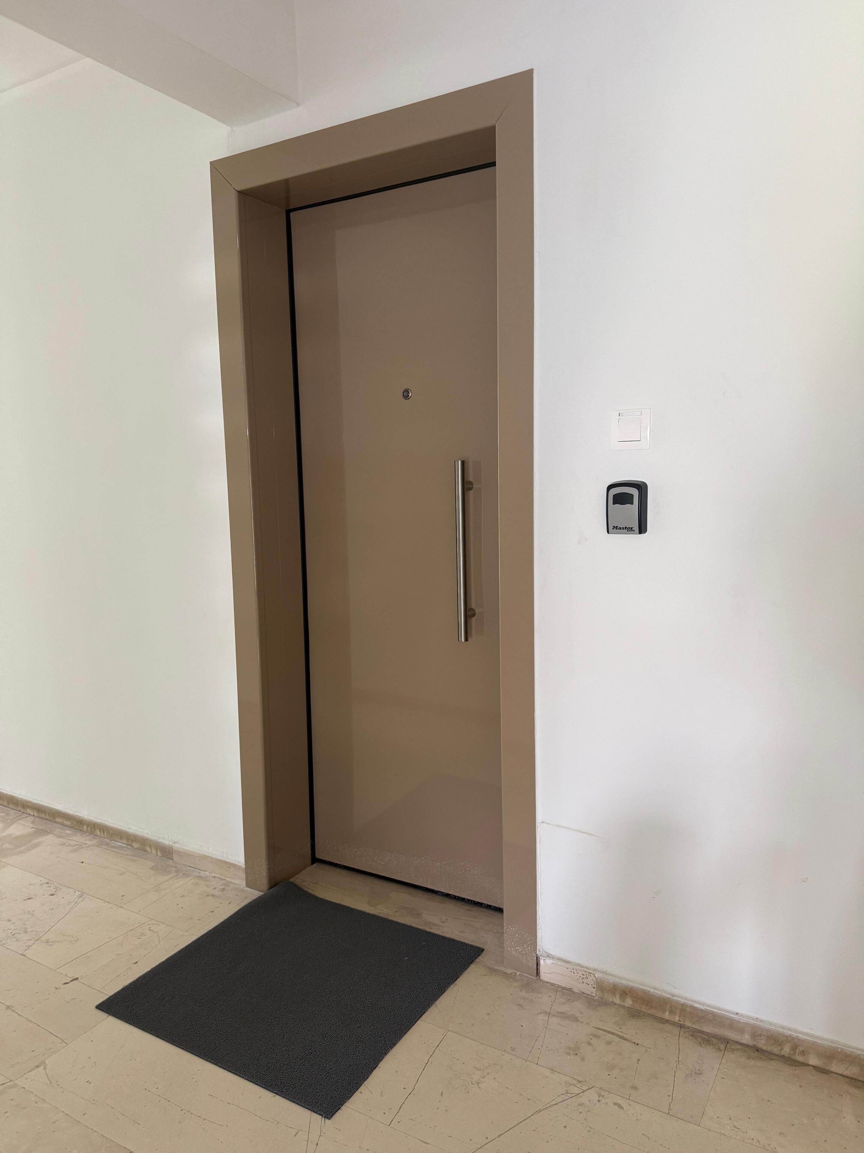 A closed door with a handle, a small electronic device on the wall, and a black doormat on the floor.