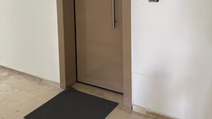 Uma porta fechada com maçaneta, um pequeno dispositivo eletrônico na parede e um tapete de entrada preto no chão.