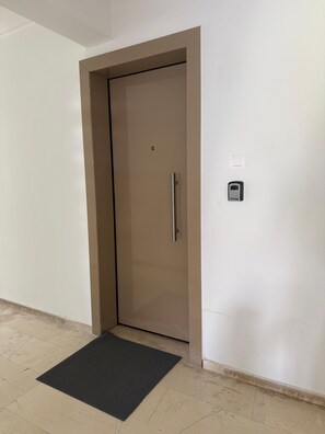 Eine geschlossene Tür mit Griff, ein kleines elektronisches Gerät an der Wand und eine schwarze Fußmatte auf dem Boden.
