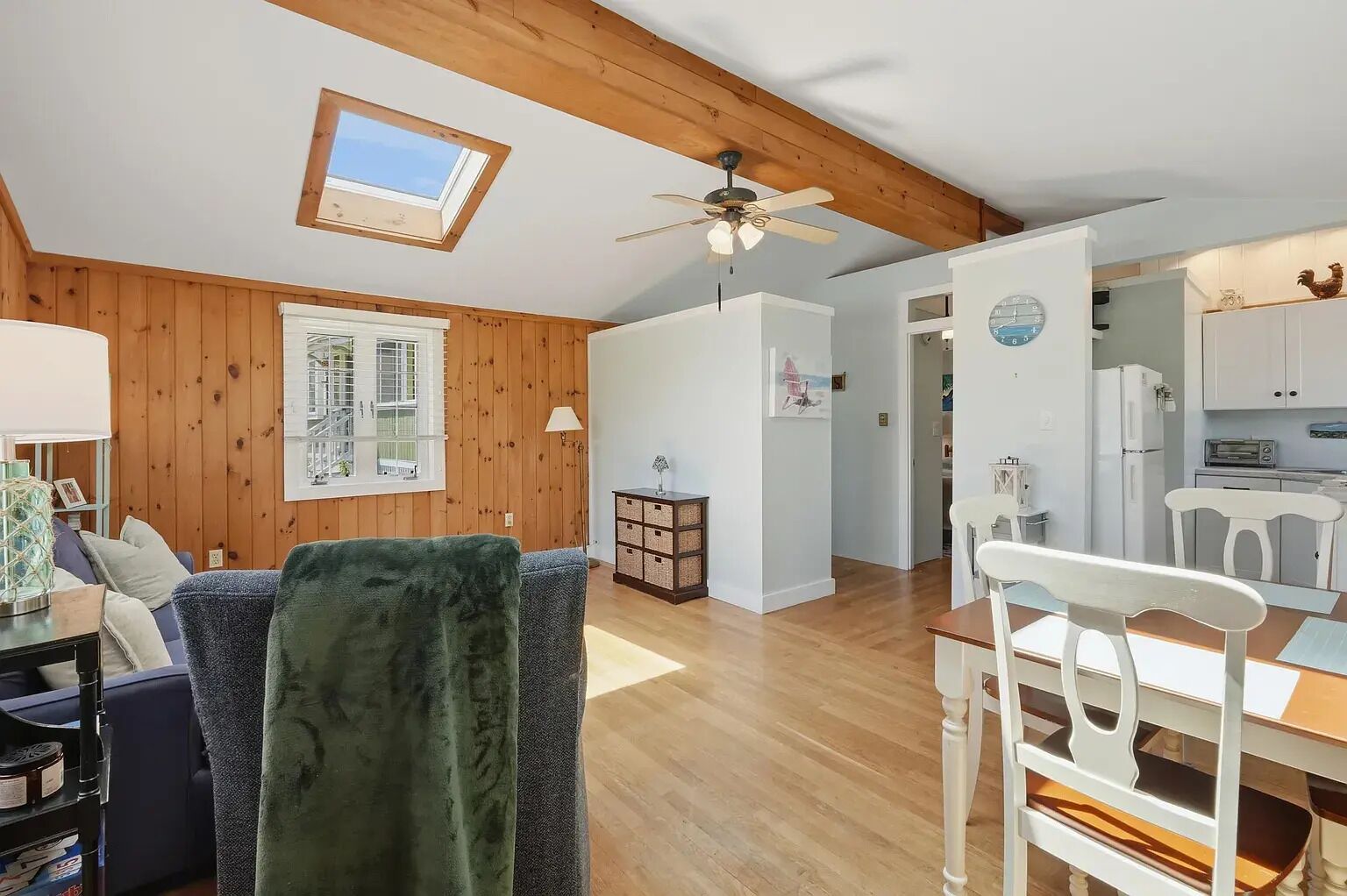 A cozy room with a wooden ceiling, a skylight, a ceiling fan, a dining table with chairs, and a kitchen area with cabinets and appliances.