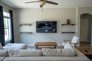A living room with a flat-screen TV mounted on the wall, a ceiling fan, a wooden coffee table, a white sofa, and a wooden chair.