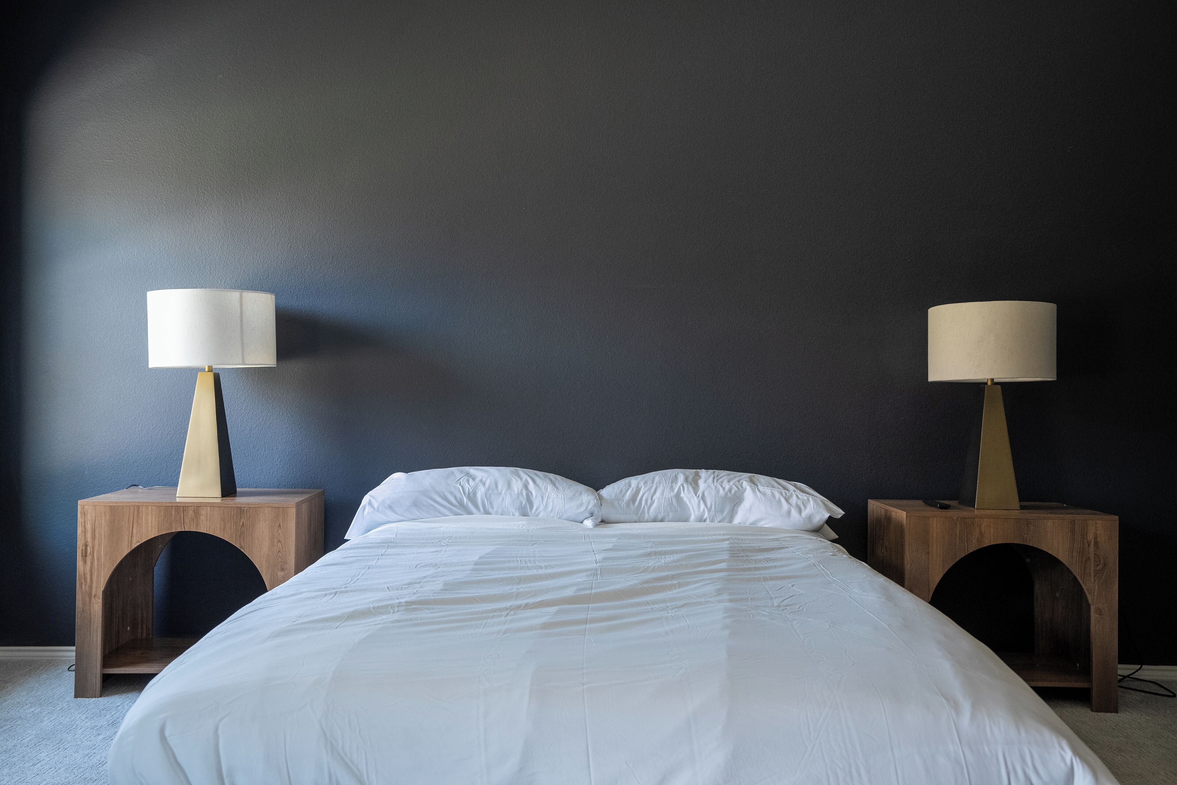 A bed with white linens, flanked by two wooden nightstands with lamps, against a dark wall.