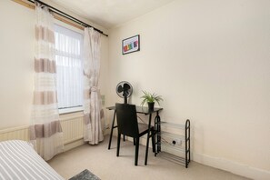 Een kamer met een bed, een bureau, een stoel, een radiator, een ventilator, een kamerplant en een foto aan de muur.