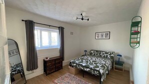Ein Schlafzimmer mit Bett, Schminktisch, einem Fenster mit Vorhängen, einem Deckenventilator und einem an der Wand befestigten Regal.