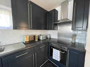 A modern kitchen with dark cabinetry, a stainless steel range hood, a built-in oven, and a countertop with a toaster and kettle. - Romford Retreat - Modern 2 Bedroom Apartment (Romford)