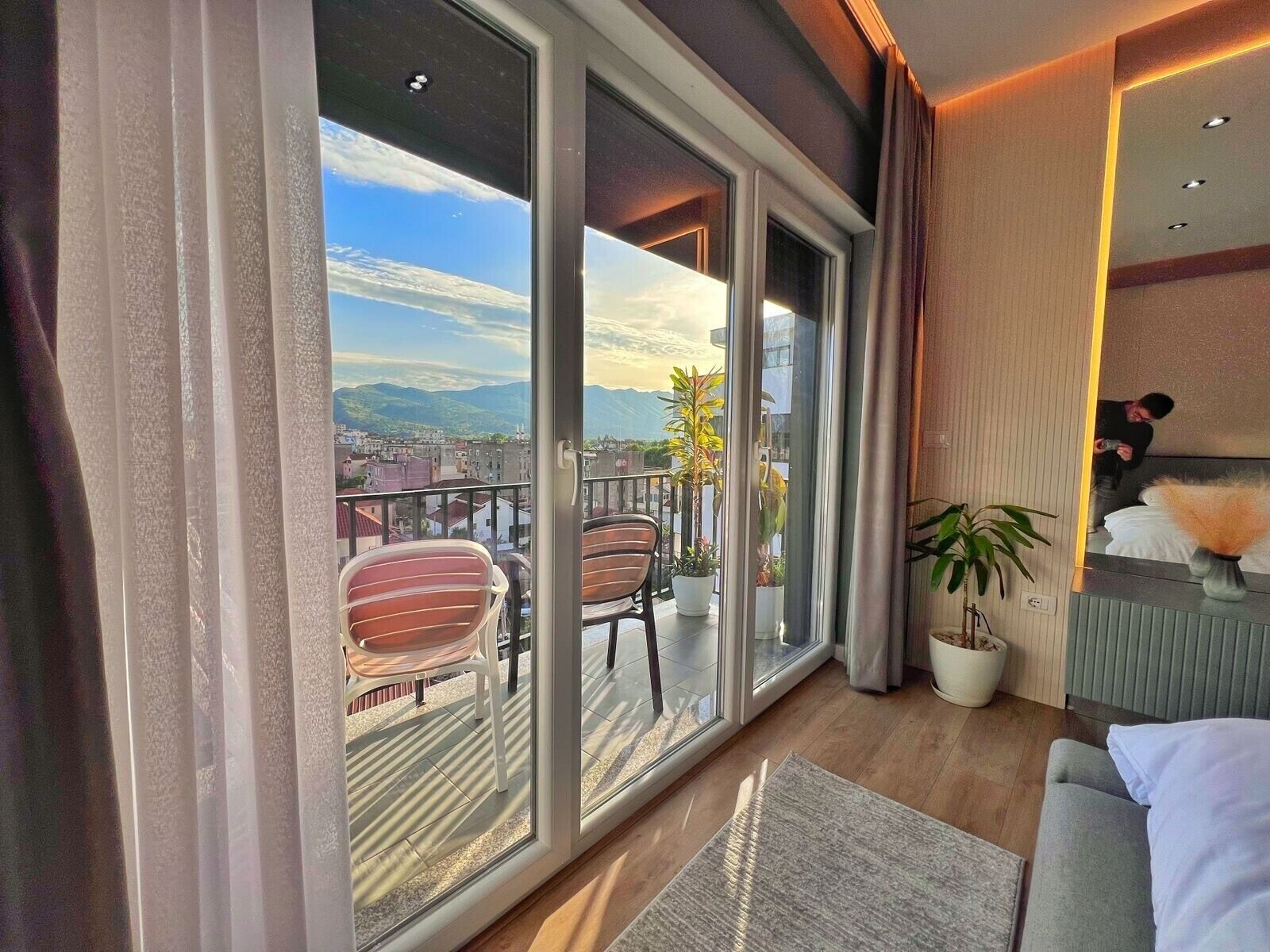 A modern hotel room with a balcony, large windows, and a view of the cityscape.