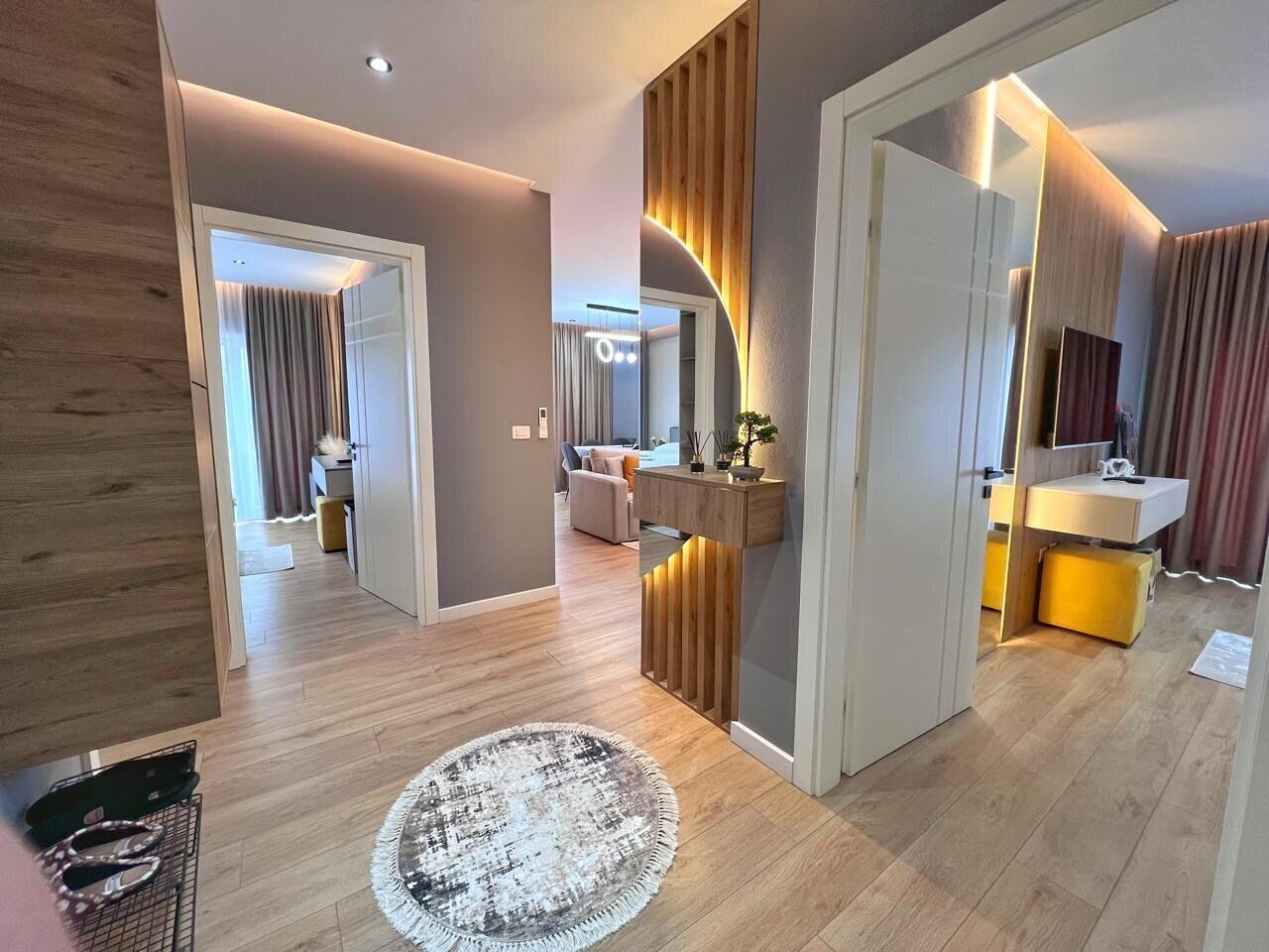 A modern interior with a wooden floor, a round rug, a wooden accent wall, a white door, and a bathroom with a yellow vanity.