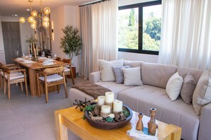 A modern living room with a wooden dining table set for four, a beige sofa with multiple cushions, and a large window with curtains.
