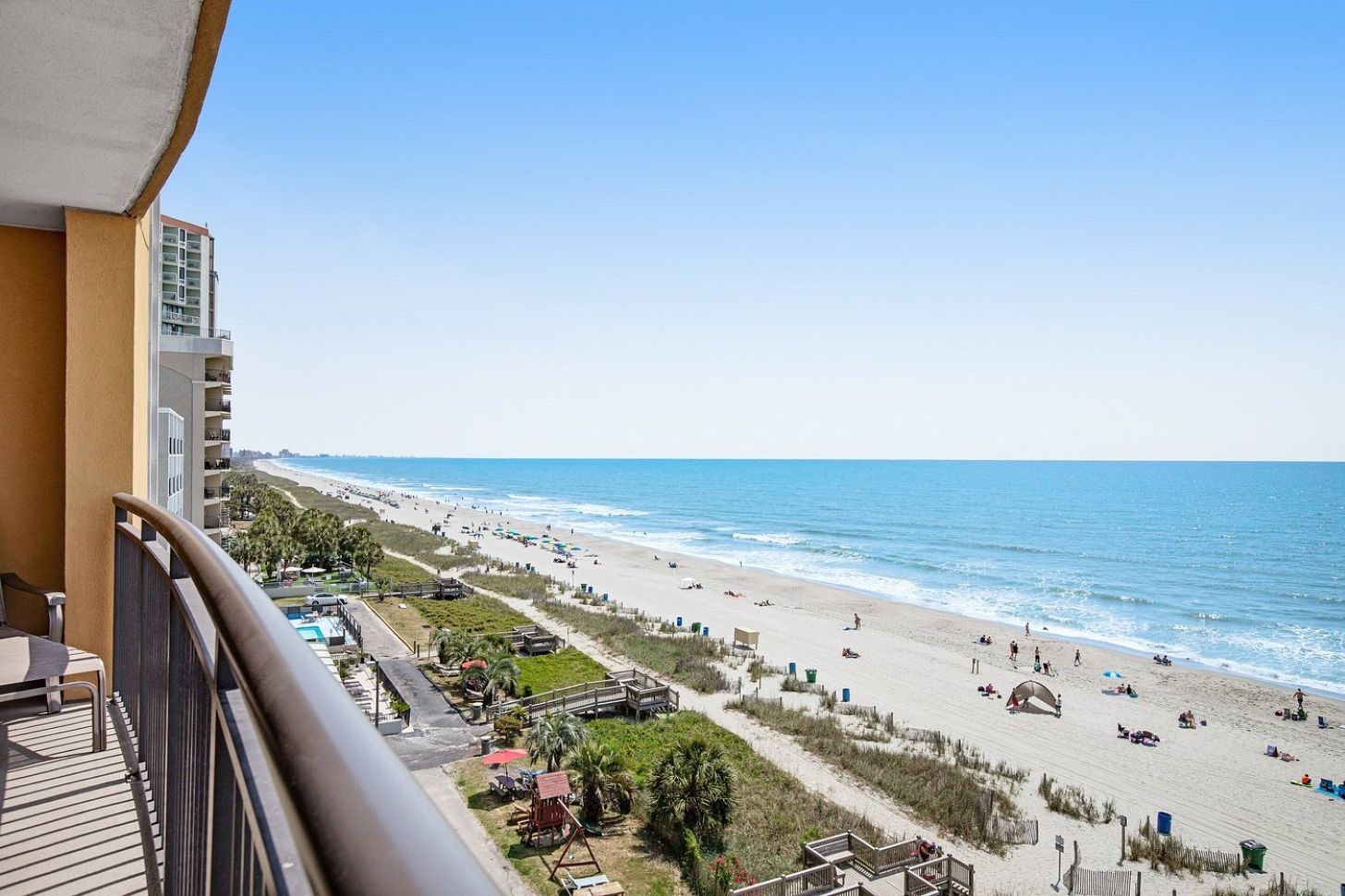 Un balcón con vistas a una playa, un área de piscina y un edificio de varios pisos.