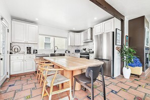 A kitchen with white cabinets, a central island with chairs, a refrigerator, and a microwave. - Charming 5BR Group Bungalow Steps to Sand with Deck and Fire Pit (Newport Beach)