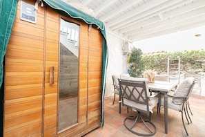 A wooden sauna with a sliding door, a table with chairs, and a view of a garden with a climbing vine. - Charming 5BR Group Bungalow Steps to Sand with Deck and Fire Pit (Newport Beach)