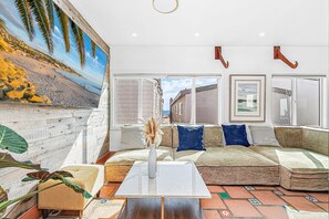 A living room with a large window, a sofa, a coffee table, and a decorative wall. - Charming 5BR Group Bungalow Steps to Sand with Deck and Fire Pit (Newport Beach)