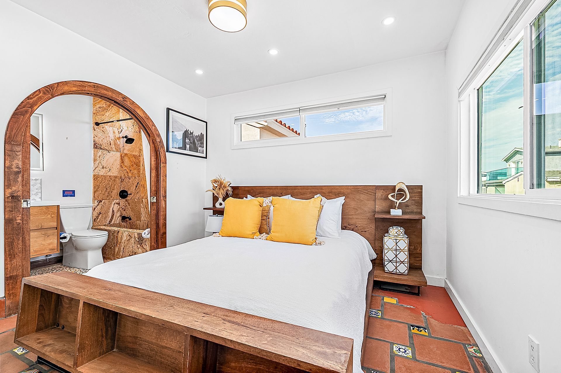 A bedroom with a large bed, wooden headboard, a wooden bench, a mirror, a window, and a view of the outside.