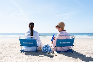 Zwei Personen sitzen auf Strandstühlen, mit Branding von 'Beachview Rentals', auf einem Sandstrand mit dem Meer im Hintergrund.