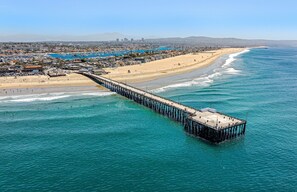 A pier extending into the ocean with a beach and cityscape in the background. - Charming 5BR Group Bungalow Steps to Sand with Deck and Fire Pit (Newport Beach)
