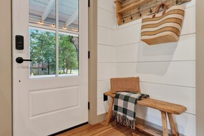 Ein Zimmer mit einer weißen Tür, einer Bank, einem gewebten Sitzkissen, einem an der Wand befestigten Regal mit einer gestreiften Tasche und Blick auf Bäume durch ein Fenster.