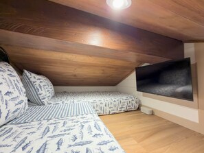 Ein Schlafzimmer im Spitzboden mit schrägen Wänden, einem großen an der Wand befestigten Fernseher und einer bettenüberdeckenden Tagesdecke mit Muster.