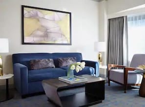 A modern hotel room with a blue sofa, a coffee table, a side table, a chair, and a large abstract painting on the wall.