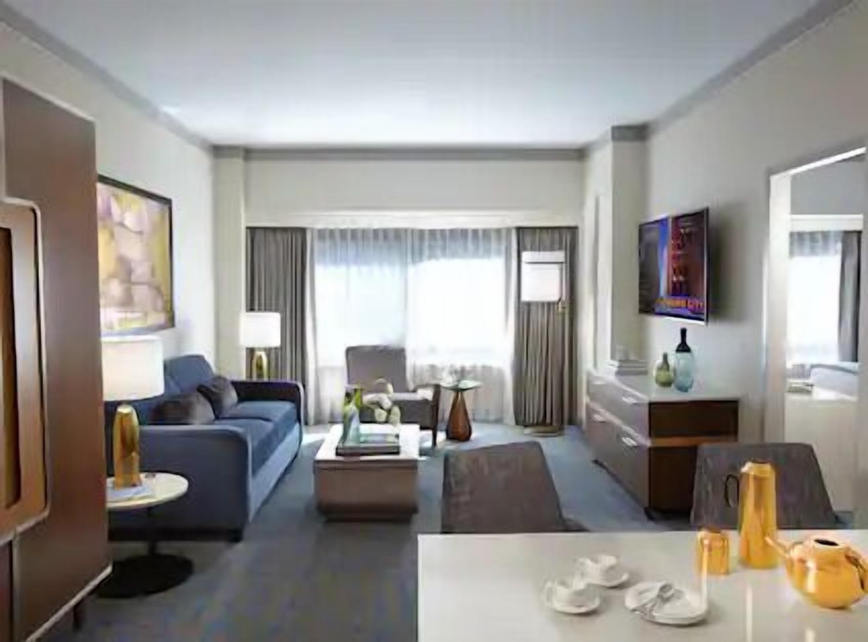 Ein Hotelzimmer mit einer Couch, einem Couchtisch, einem Fernseher, einem Schreibtisch und einem Esstisch mit Schuhen und einem Wasserkocher.