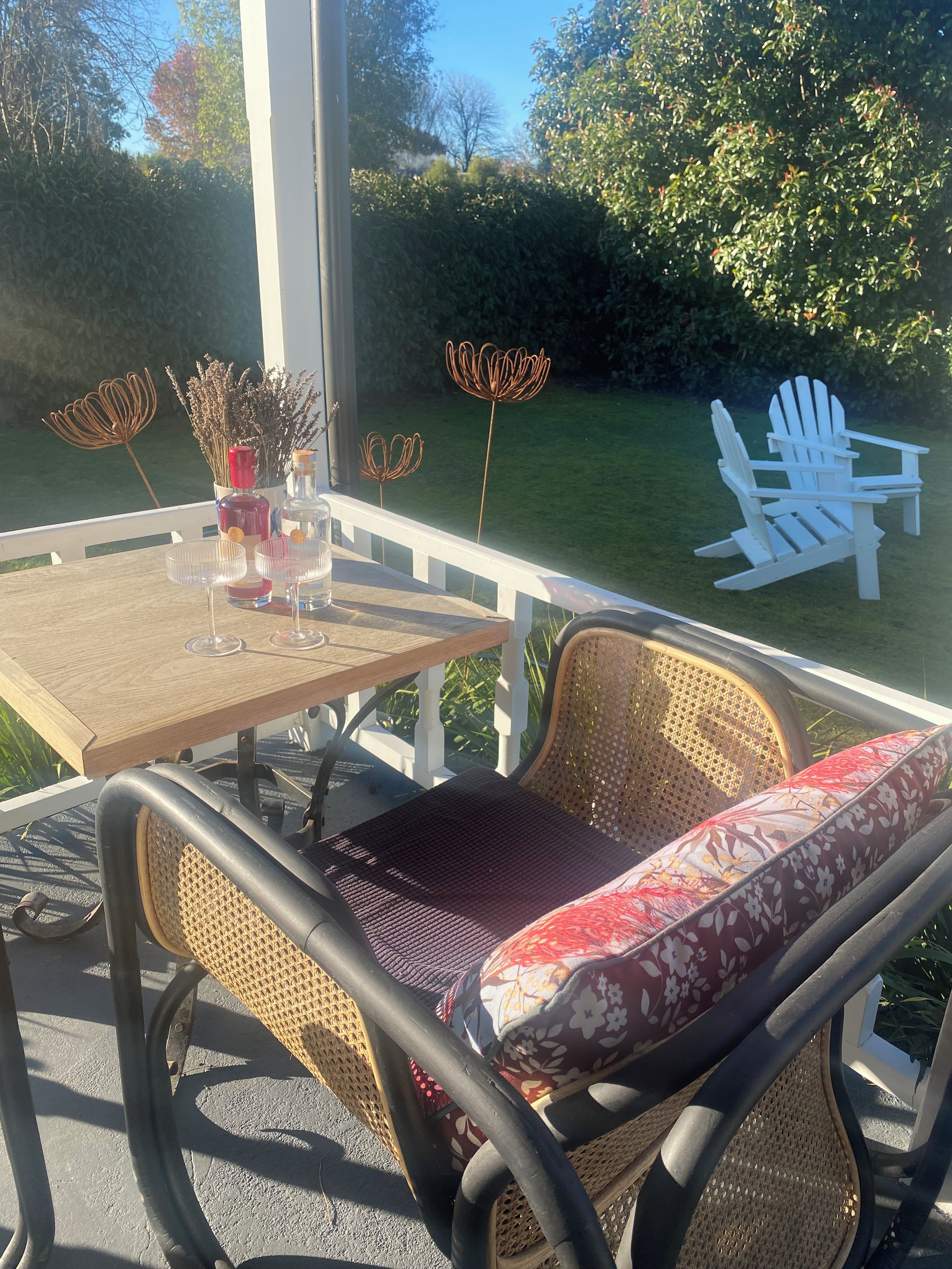 A patio with a table set for two, a wicker chair with a cushion, and a white Adirondack chair.