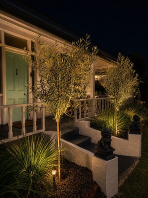 A house with a light green door, a porch with a railing, and a well-lit garden with various plants and a statue.