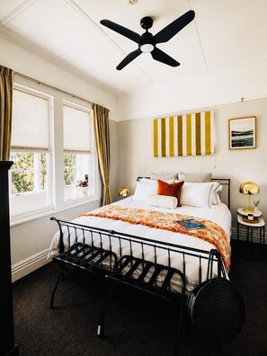 A bedroom with a bed, a ceiling fan, a window with curtains, a bedside table with a lamp, and a wall with a striped artwork.