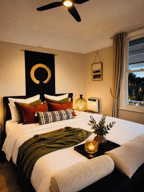 Ein Schlafzimmer mit einem Bett, Deckenventilator, Nachttisch, Lampe, einem Fenster mit Vorhängen und einem Wandbild.